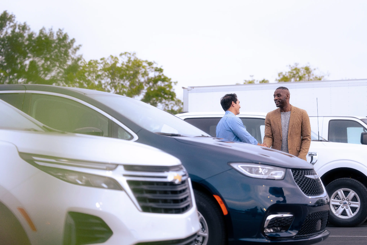 Two smiling men shaking hands next to a minivan in a car sales lot.