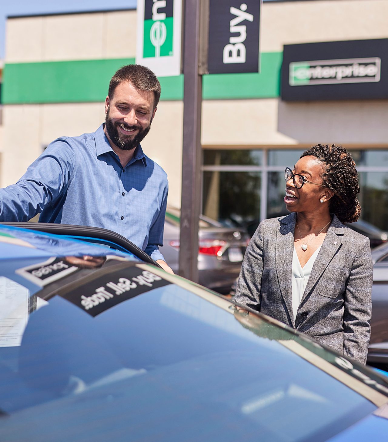 A gentleman, standing in front of a blue car, speaks with a lady at the Enterprise Car Sales lot.
