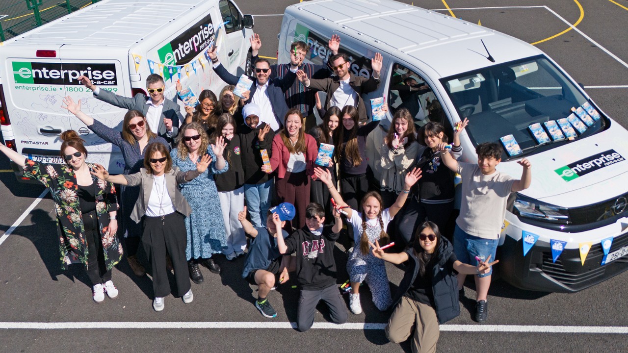 Sevel adults and children, flanked by two Enterprise Rent-A-Car vehicles, hold up books in a parking lot on a sunny day