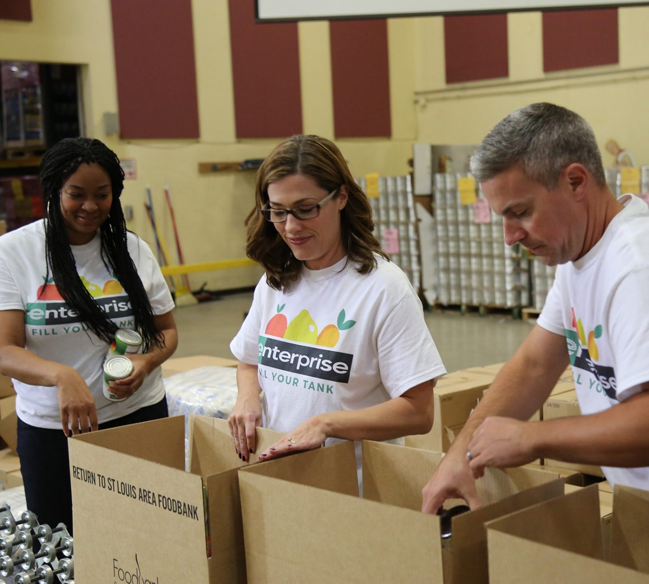 Two female Enterprise employees and one male employee, all wearing white Enterprise Fill Your Tank t-shirts, pack boxes during a community service project.