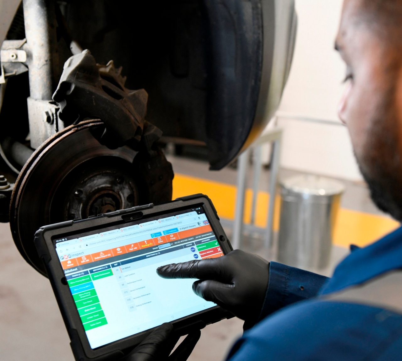 A male Enterprise employee, wearing a black glove in an auto mechanic's garage, points at an iPad.