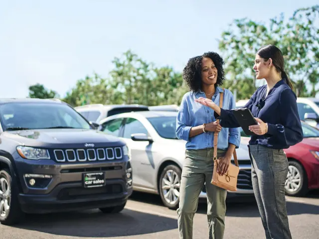 Dos mujeres hablando entre sí frente a un lote de ventas de autos.